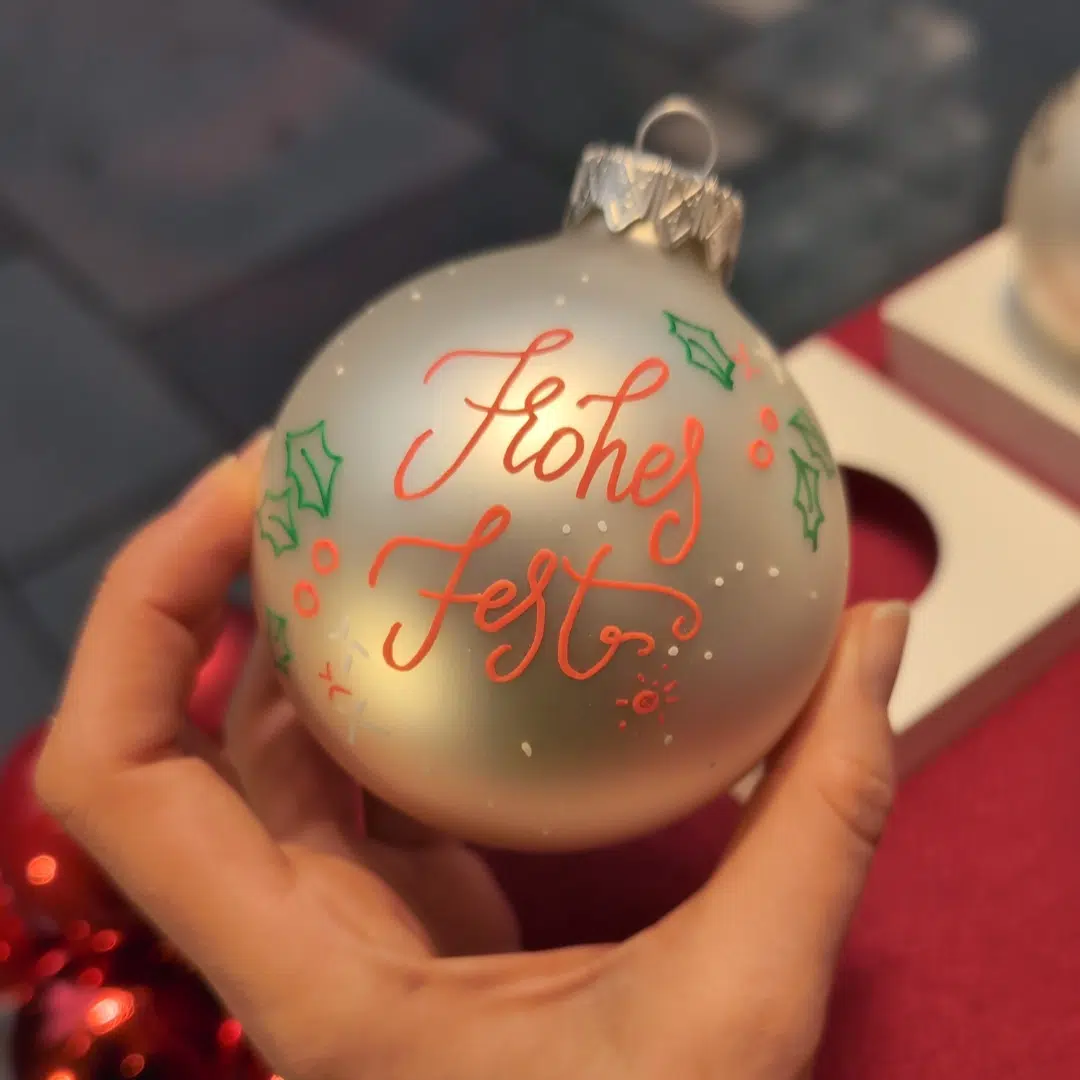 Christbaumkugel Lettering: Eine Frau hält eine goldene Christbaumkugel in der Hand, auf der in roter Handlettering Schrift "Frohes Fest" steht.